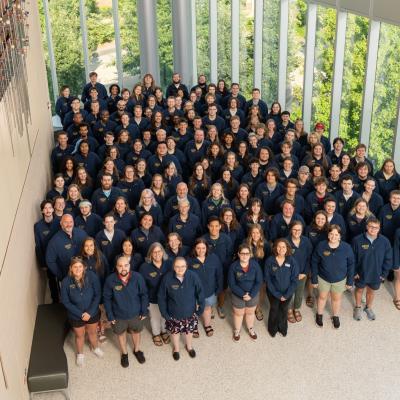 A large group of people in matching navy blue jackets poses for a group photo in a bright indoor space with tall windows and greenery outside.